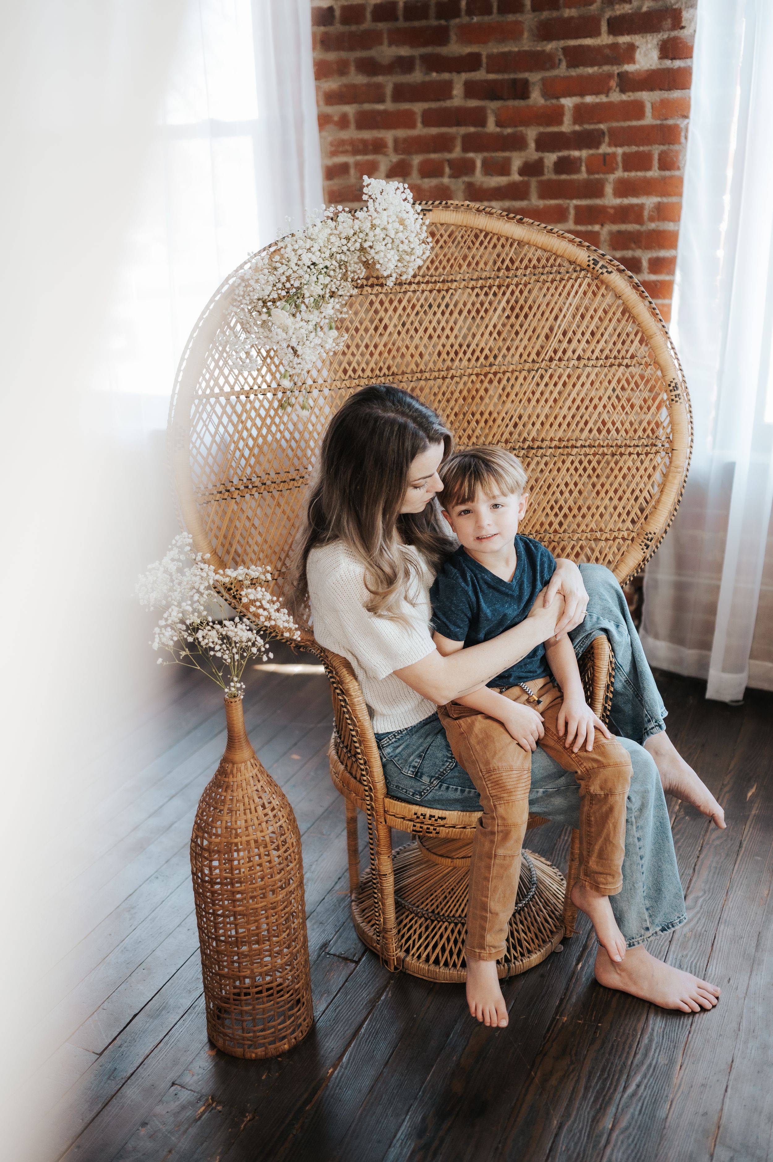 Two people sitting together in a woven peacock chair against exposed brick wall with white curtains and dried flowers.