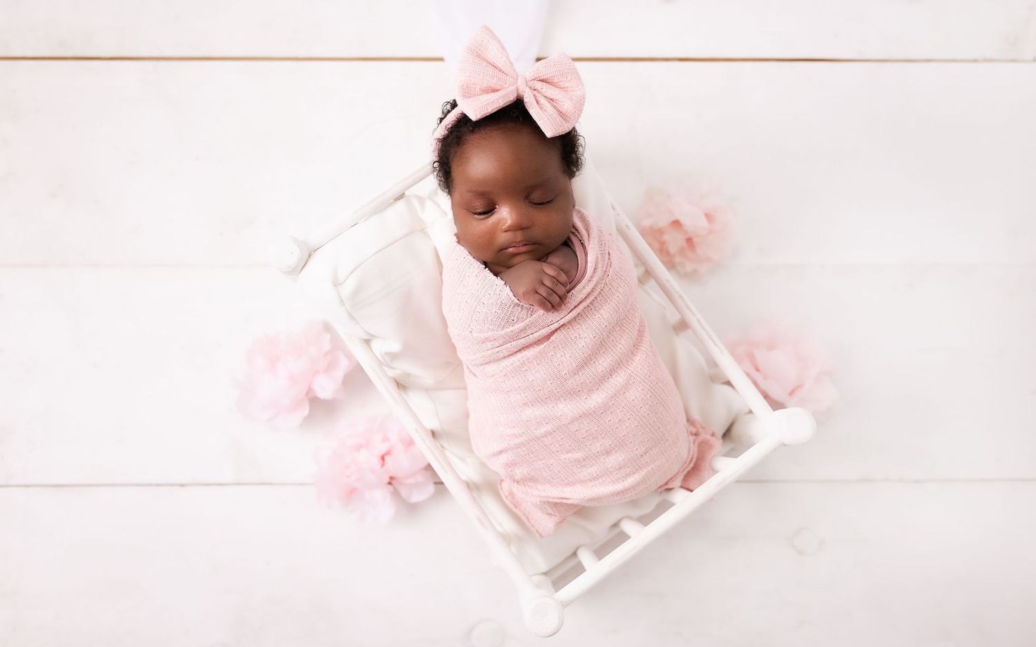 A baby wrapped in soft pink swaddle lies in a white crib surrounded by delicate pink bows.