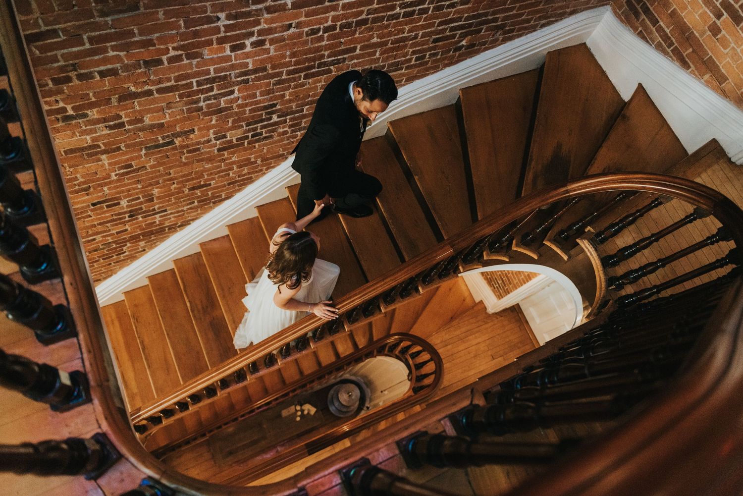 Person descending a spiral staircase with wooden steps and exposed brick walls viewed from above in warm lighting.