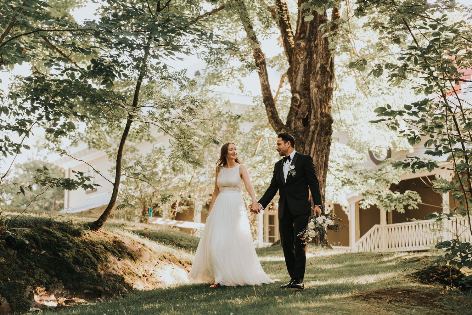 A couple walks hand in hand through a sunlit garden path with tall trees and a white picket fence in the background.