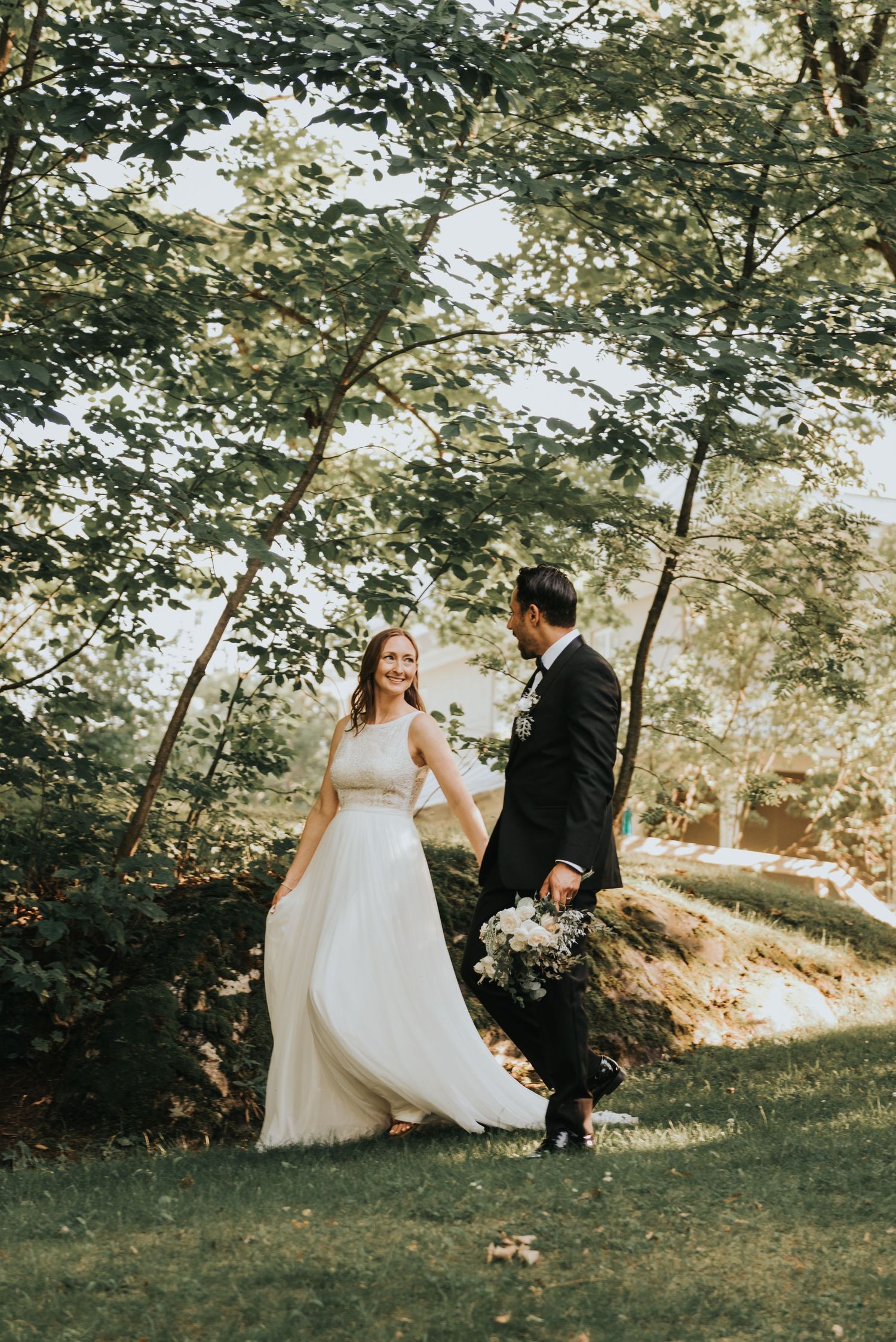 A couple in wedding attire dances playfully in a sunlit garden setting with trees in the background.
