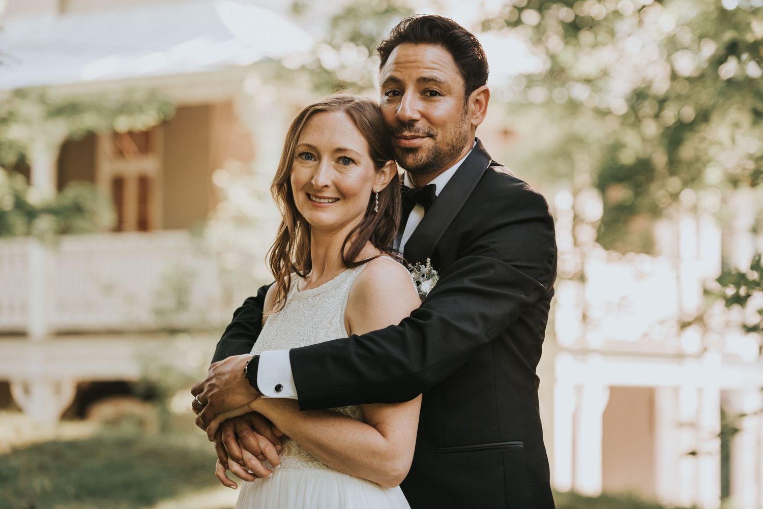 A newlywed couple embraces outdoors on their wedding day in front of a light colored building.