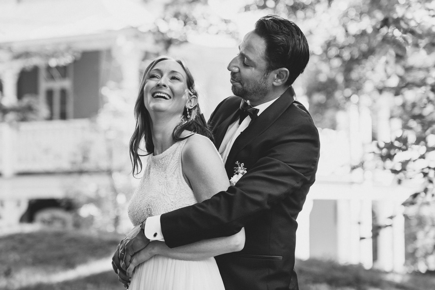 A joyful black and white photograph of newlyweds sharing a laughing embrace during their outdoor wedding celebration.