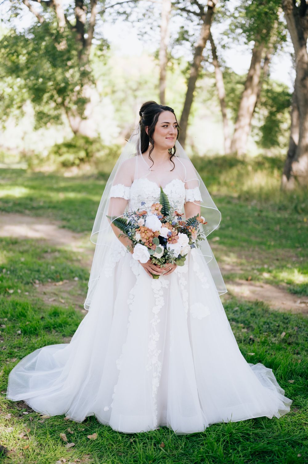 Two brides in white wedding dresses sit together on grass holding floral bouquets in a sunlit outdoor setting.