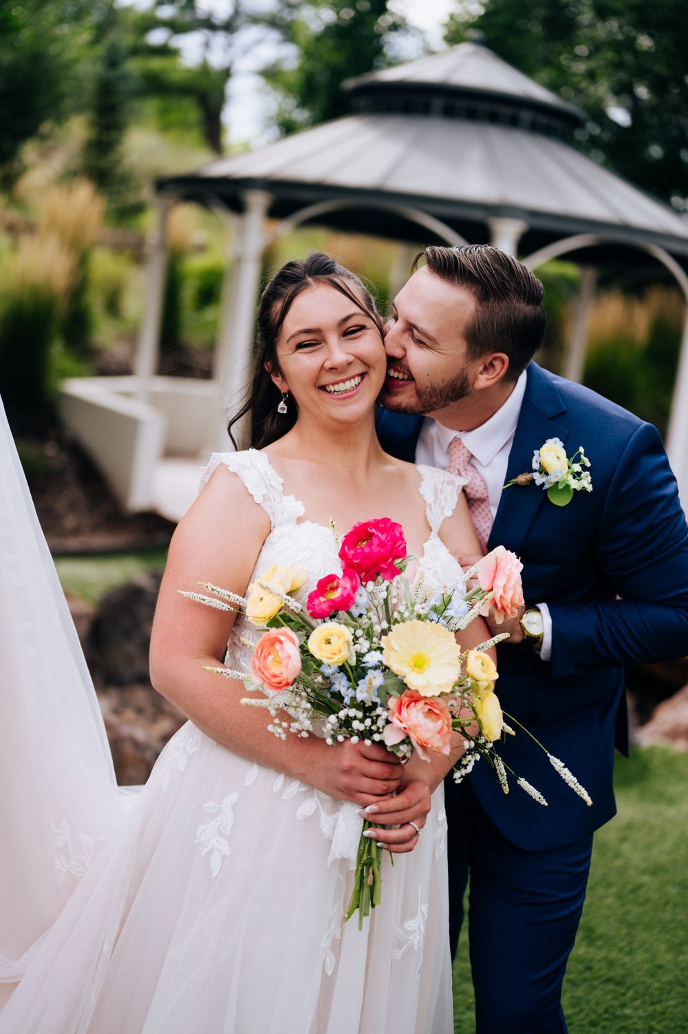 A romantic wedding couple embraces outdoors while holding a vibrant bouquet of pink and yellow flowers.