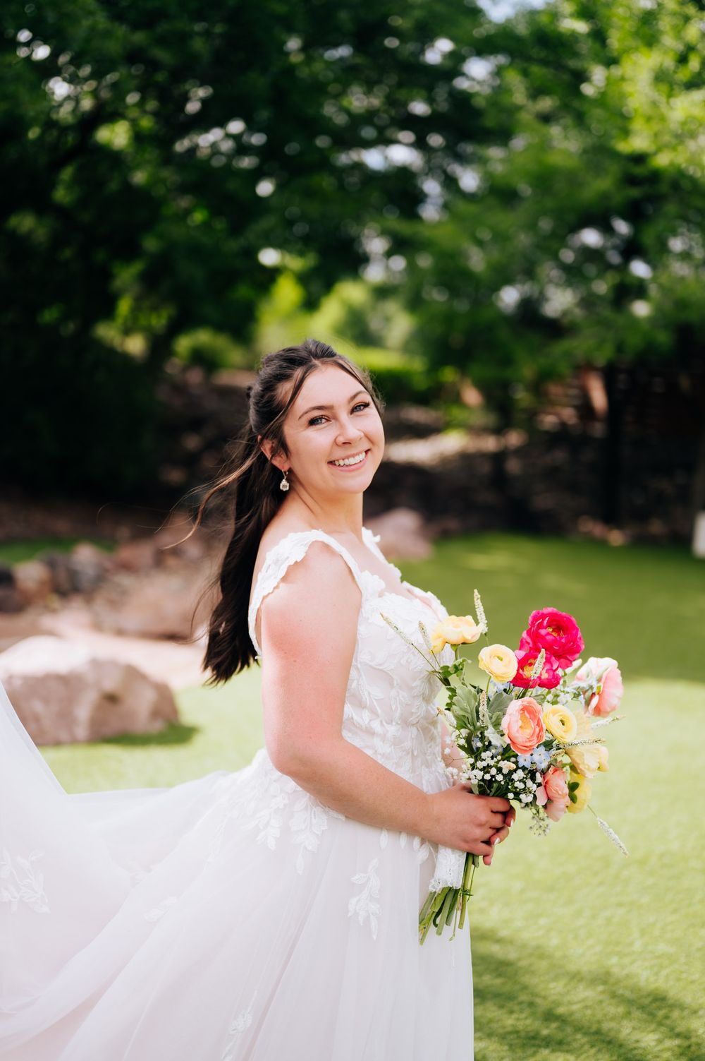 A bride in a white lace wedding dress twirls joyfully in a sunny garden setting while holding a vibrant coral bouquet.