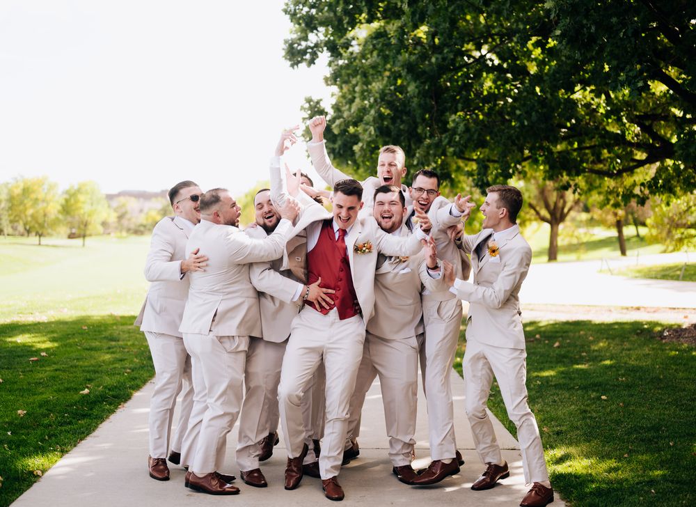 Groomsmen in beige suits playfully pose together on a sunny day in an outdoor setting.