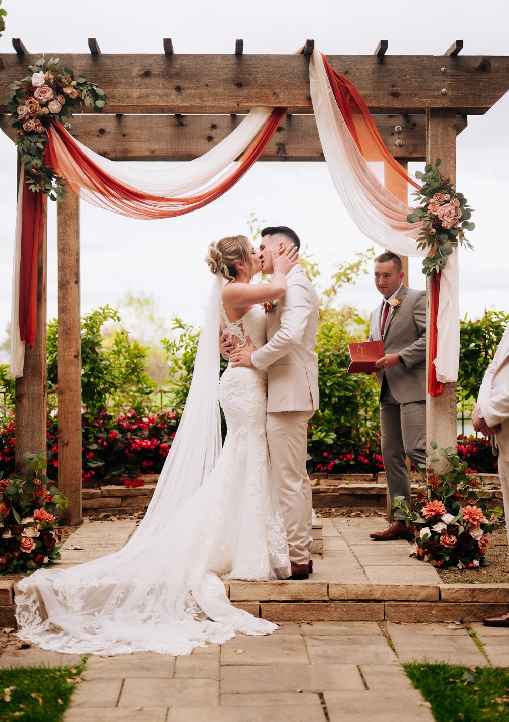 A romantic wedding ceremony under a wooden pergola decorated with coral drapes and floral arrangements on a stone patio.