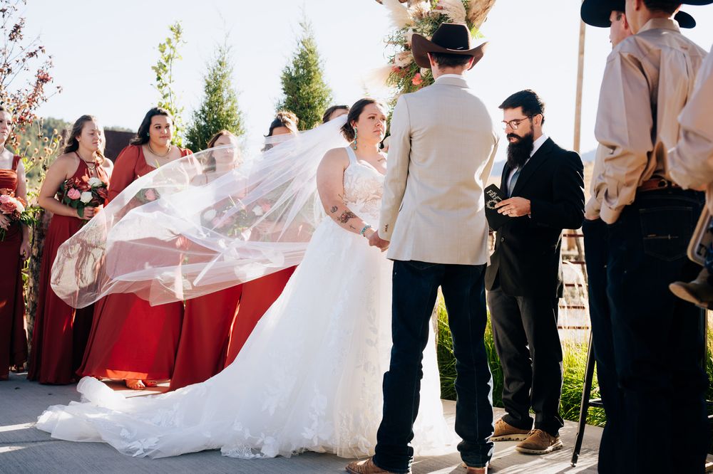 A flowing white wedding dress veil billows in the wind as a couple stands together during an outdoor ceremony.