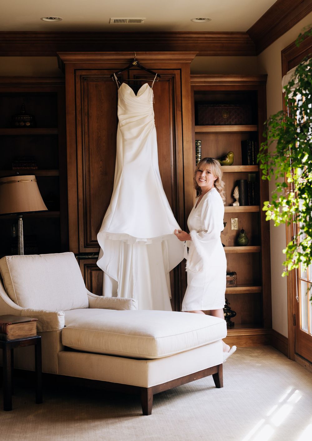 A white wedding dress hangs elegantly in a luxurious dressing room with warm wooden paneling and natural light.