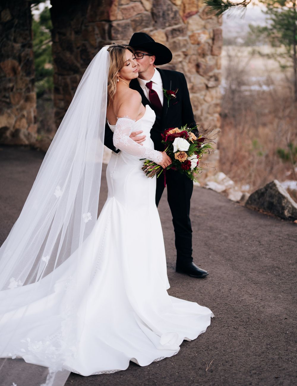 A romantic wedding couple embraces on a rustic stone path as the bride's flowing white veil catches the breeze.