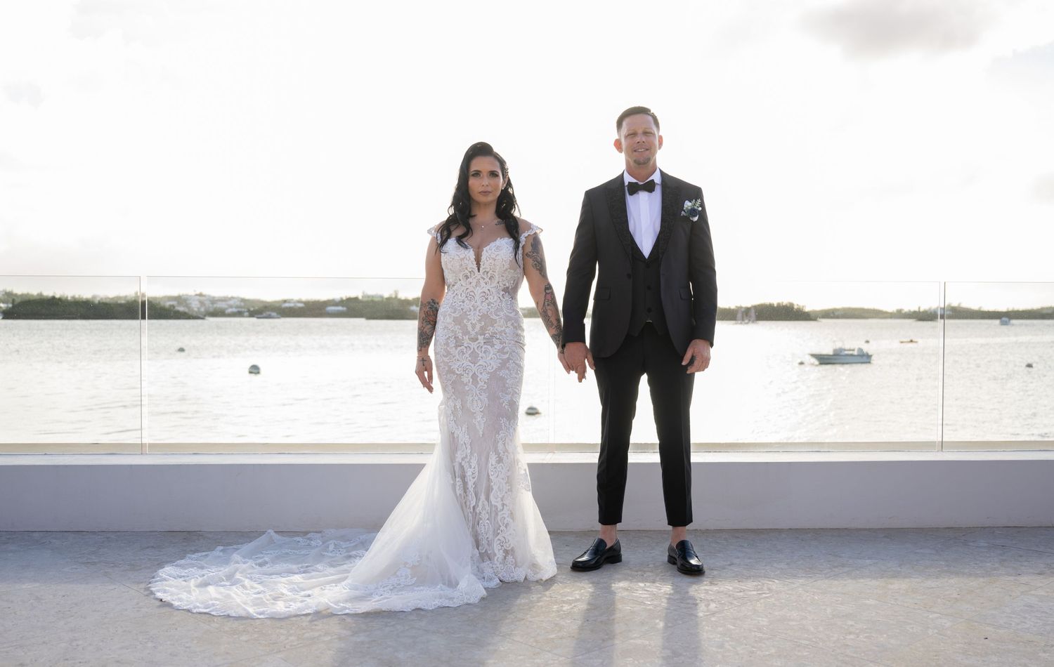 A bride and groom pose together on a white balcony with boats visible in the background.