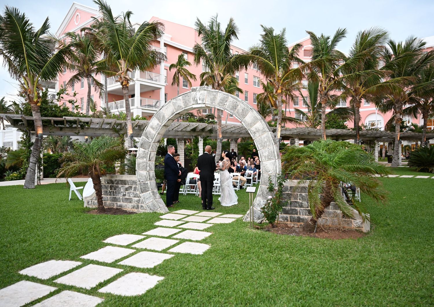 An outdoor wedding ceremony setup with a stone arch and pink resort building in the tropical background.
