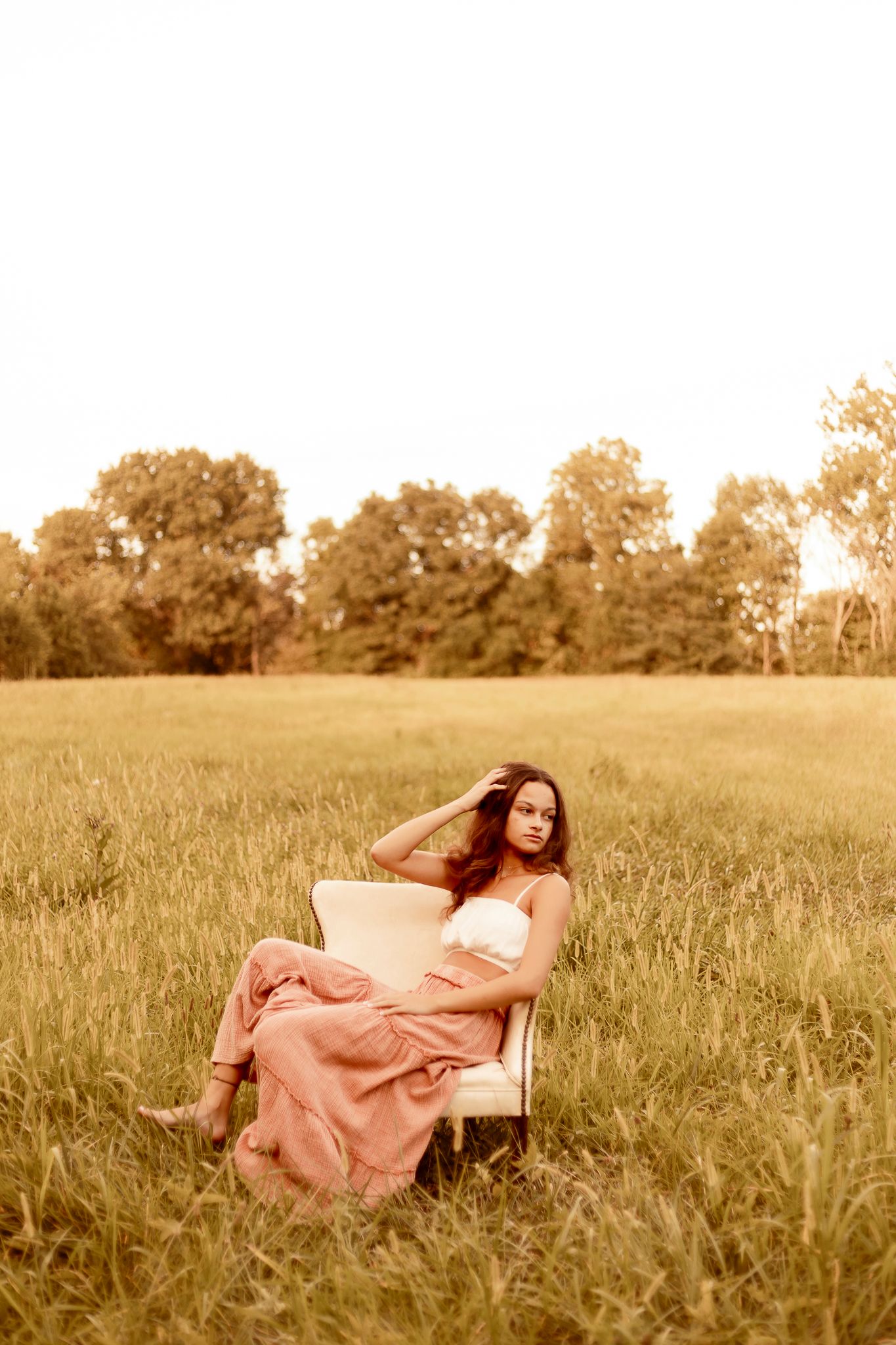 A dreamy vintage-style portrait of someone reclining in a white chair amidst a golden field at sunset.