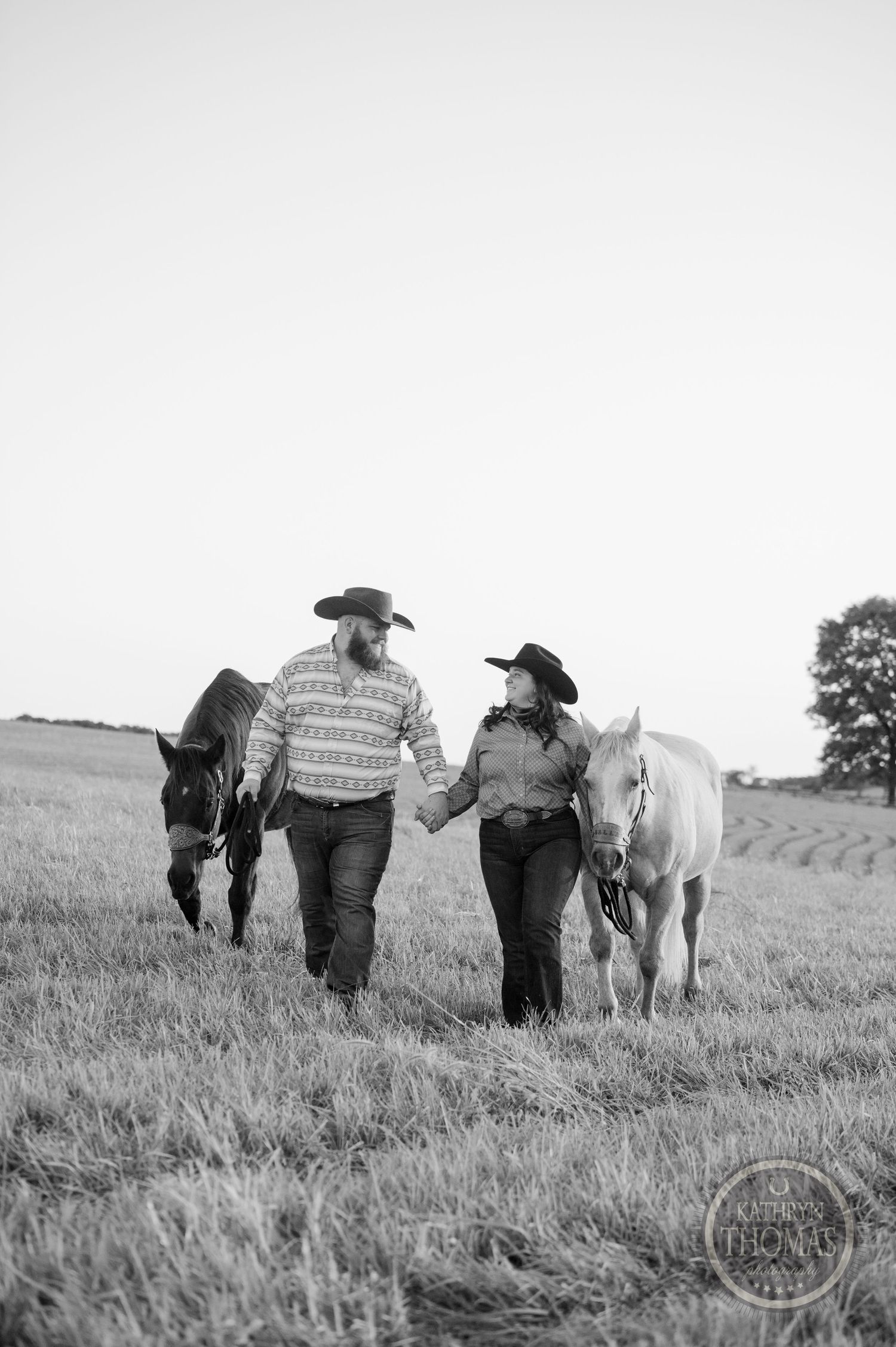 A couple in western attire walking with two horses through a grassy field in black and white.
