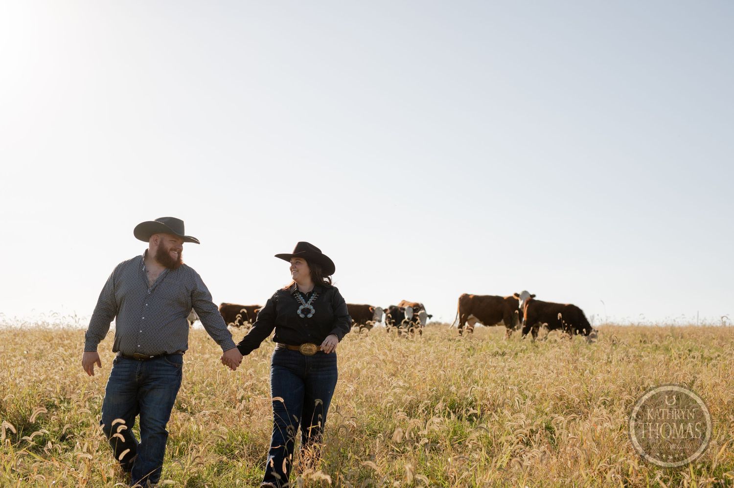 Couple holding hands walking through prairie field with cattle grazing in background.