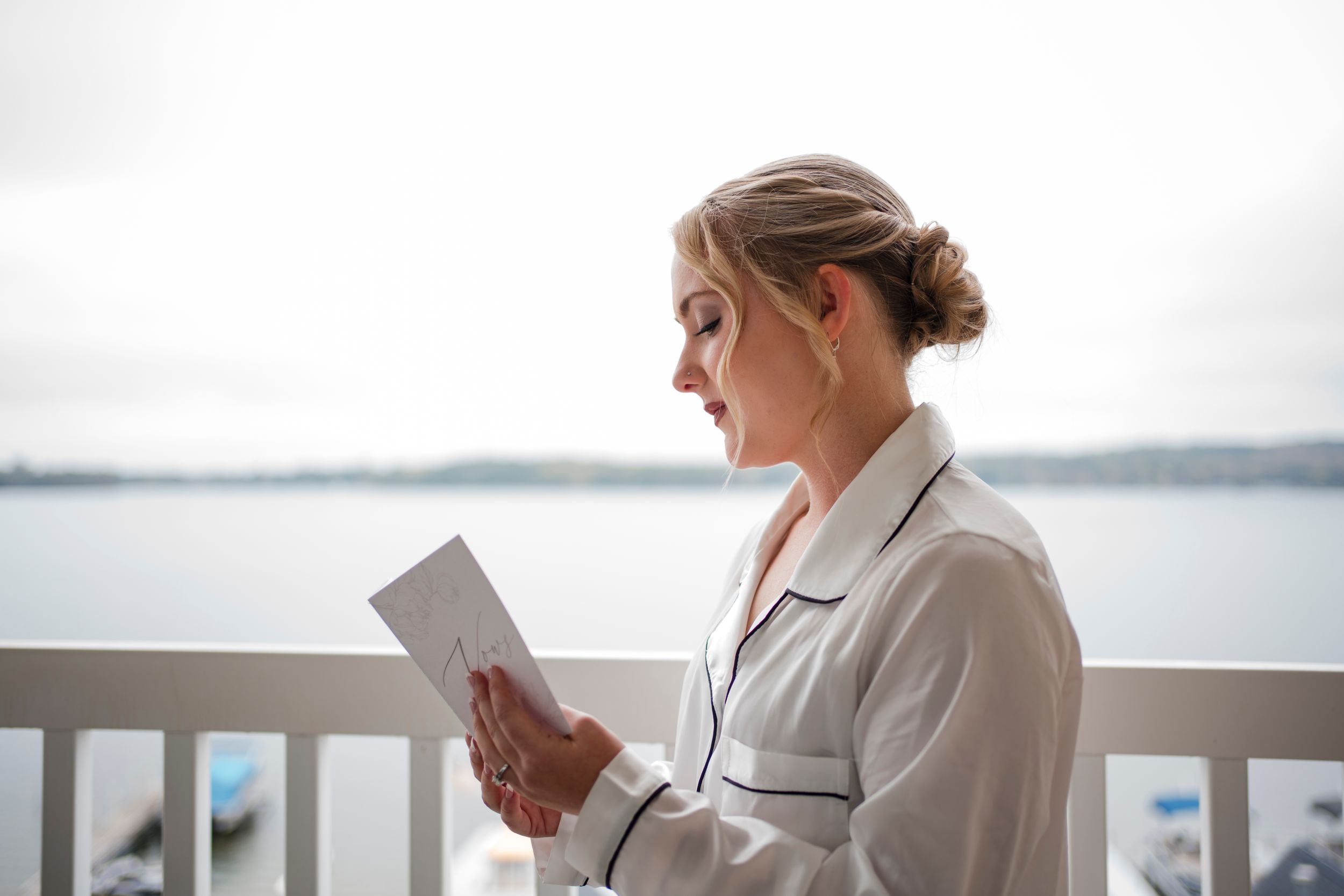 A woman in a white robe reads a letter on a balcony overlooking water.