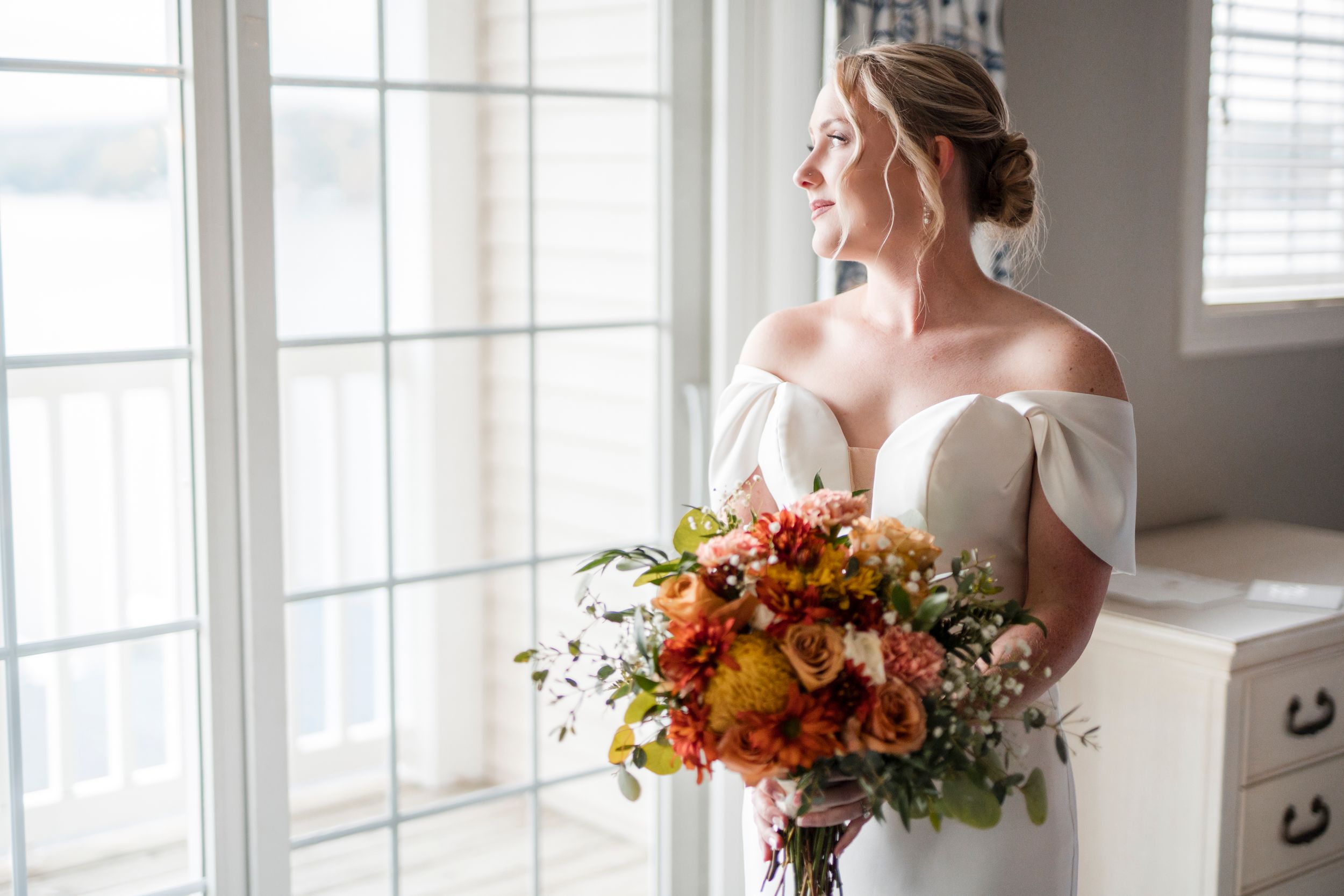 A bride holds a vibrant fall-colored bouquet while standing by window in an off-shoulder wedding dress.