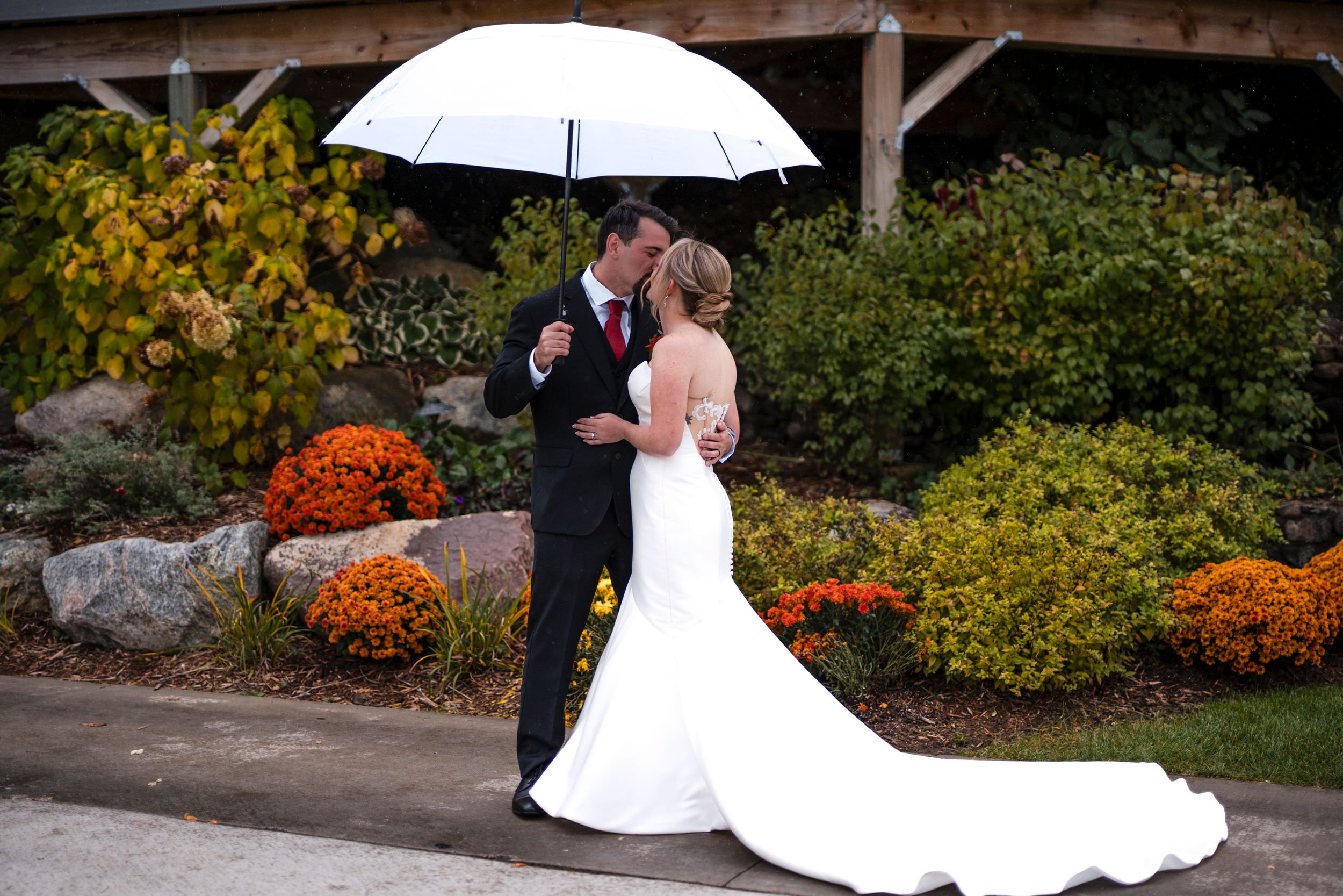 A couple shares a romantic kiss under a white umbrella in a garden with fall colors.