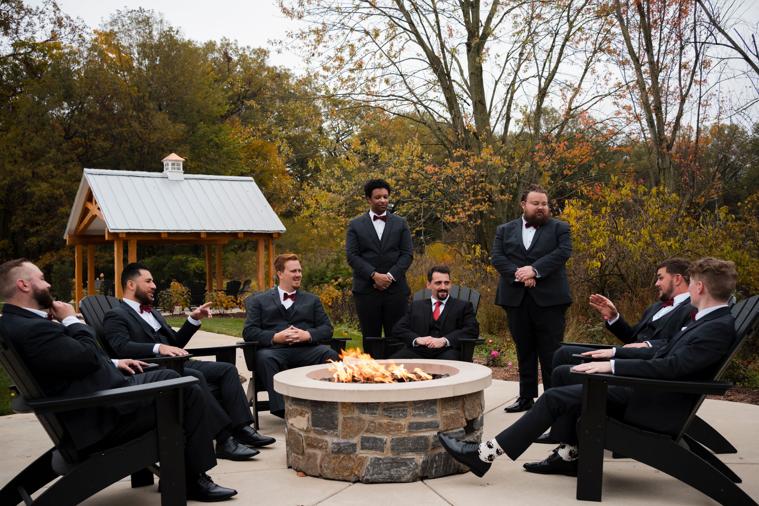 Groomsmen gather around a stone fire pit in formal attire during an outdoor wedding celebration.