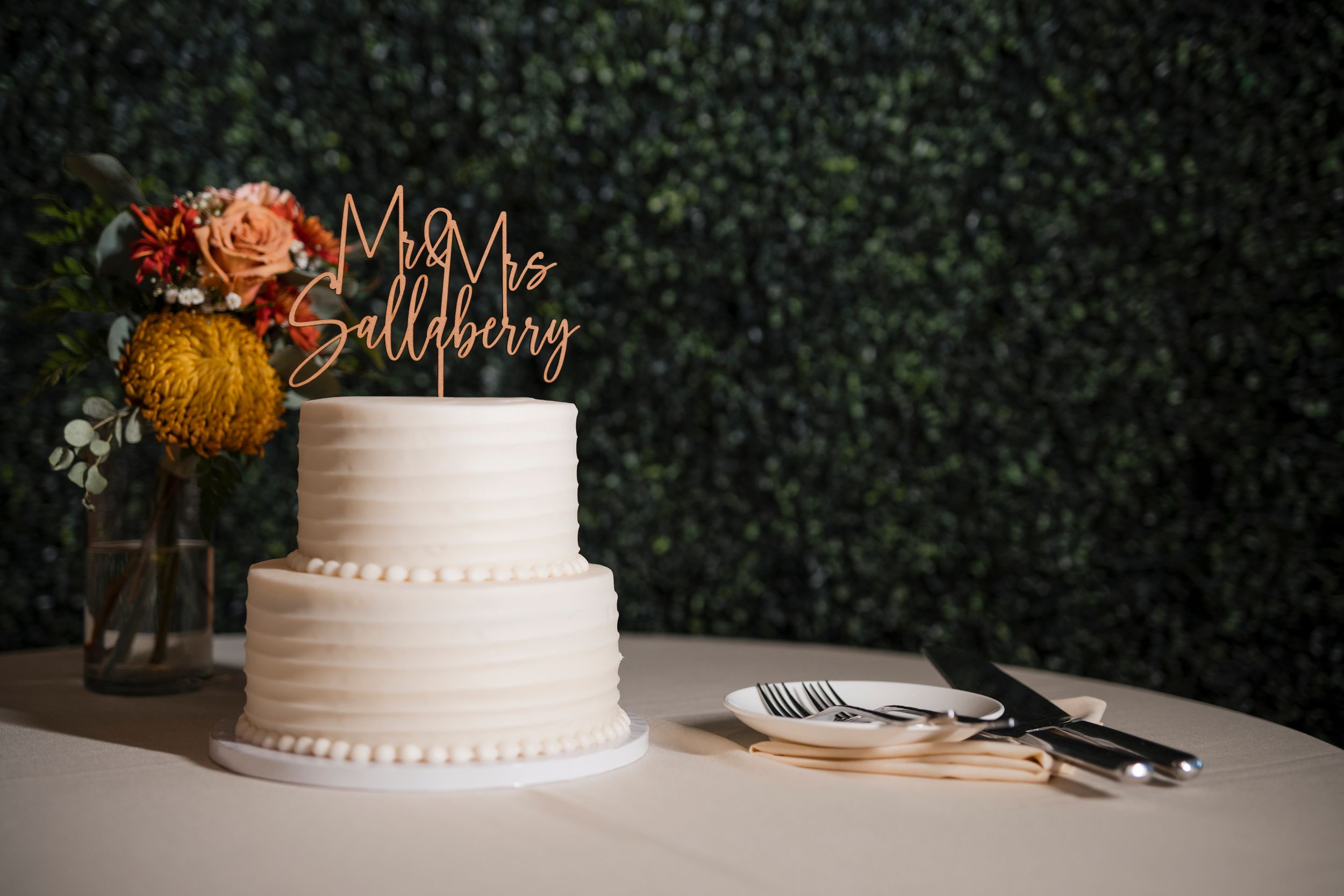 An elegant two-tier white wedding cake with floral decorations sits on a table against a dark green backdrop.