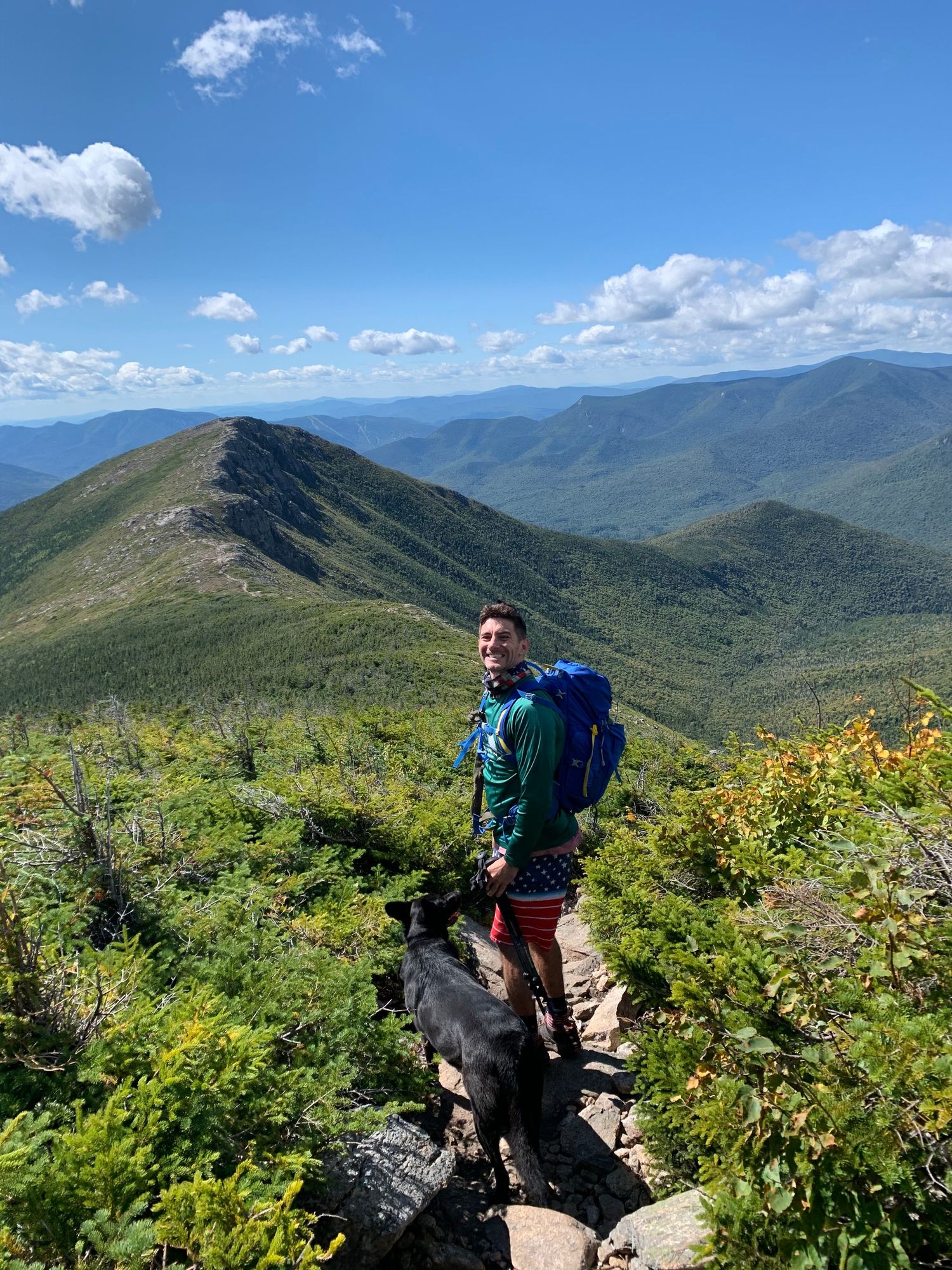Hiker on mountain trail overlooking green valley and distant peaks.