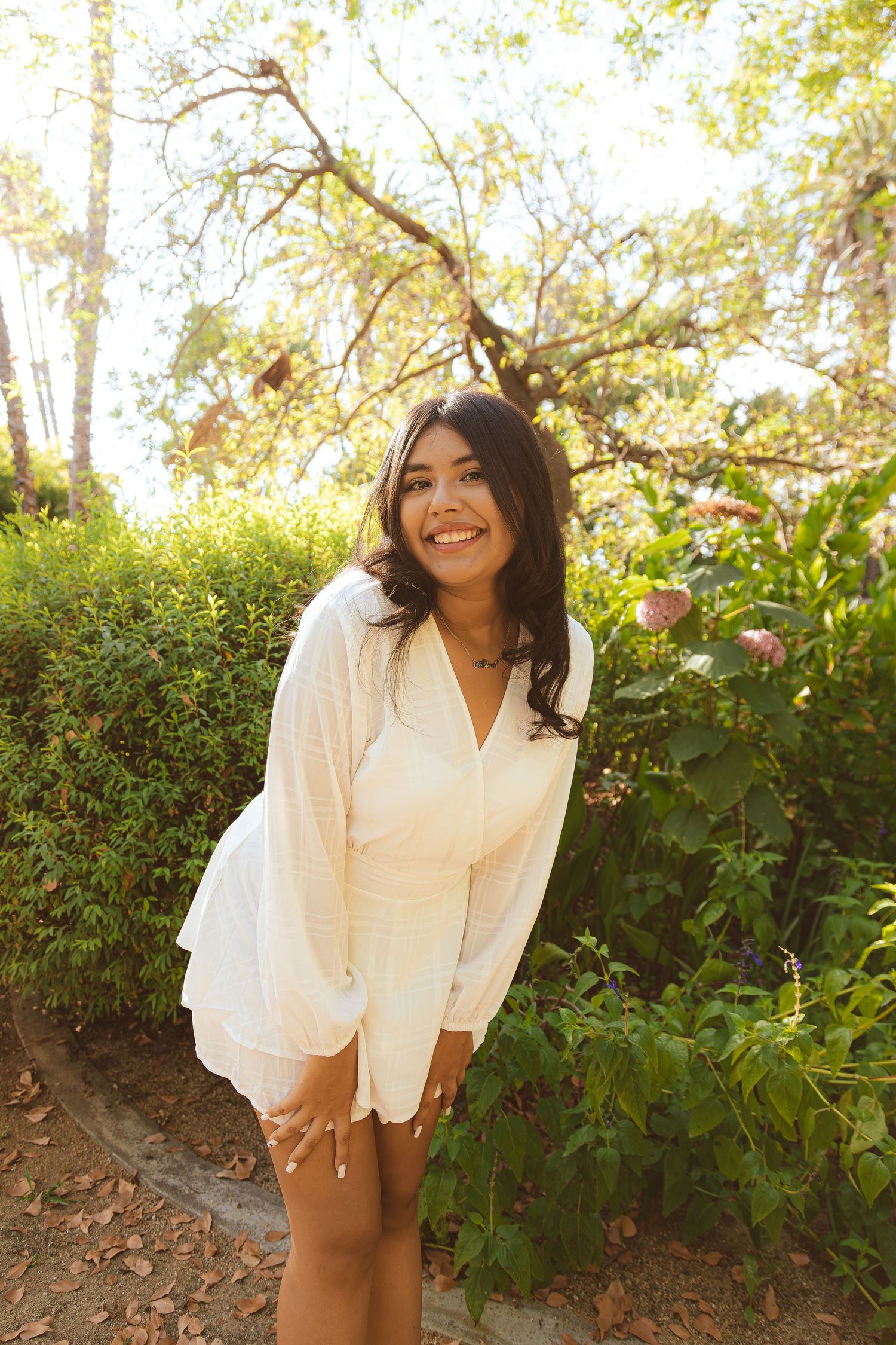 A woman in a white dress stands along a garden path on a sunny day, surrounded by lush greenery and flowers.