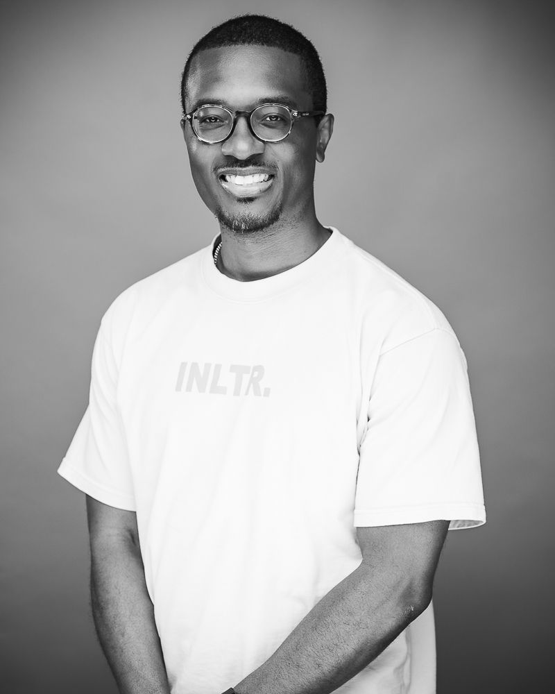 A black and white portrait of someone wearing glasses and a white t-shirt while smiling warmly against a neutral background.