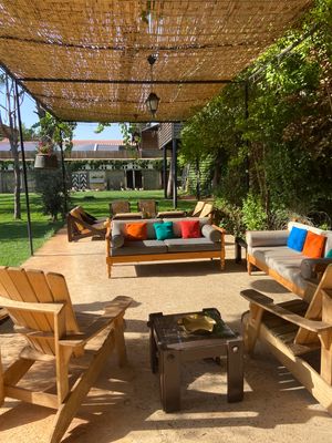 Cozy outdoor patio space with wooden Adirondack chairs, bamboo shade cover, and colorful throw pillows on wooden benches.