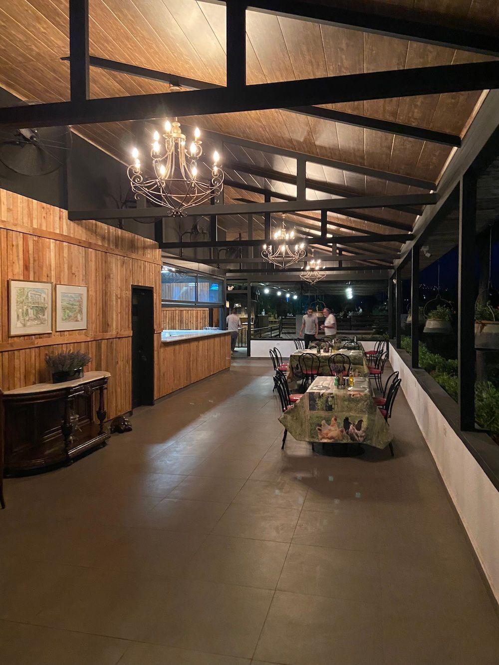Modern rustic interior hallway with exposed wooden beams, chandeliers and large windows overlooking a nighttime view.