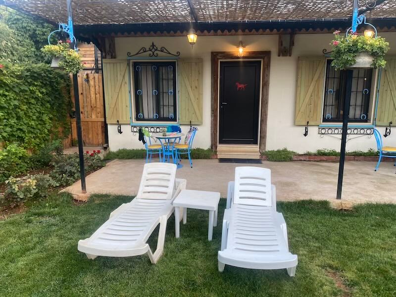 Two white patio lounge chairs sit on grass in front of a cream-colored house with dark shutters and hanging plants.