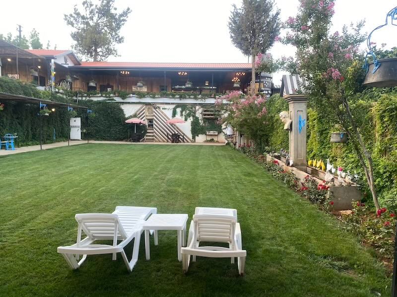 Two white lounge chairs sit on a manicured lawn in front of a terraced garden and house with covered patio.