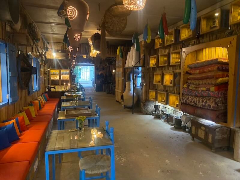 Rustic restaurant interior with blue tables, orange bench seating, and vintage decor illuminated by ambient lighting.