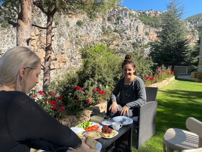 Two people enjoying an outdoor meal on a patio with dramatic rocky cliffs and colorful flowers in the background.