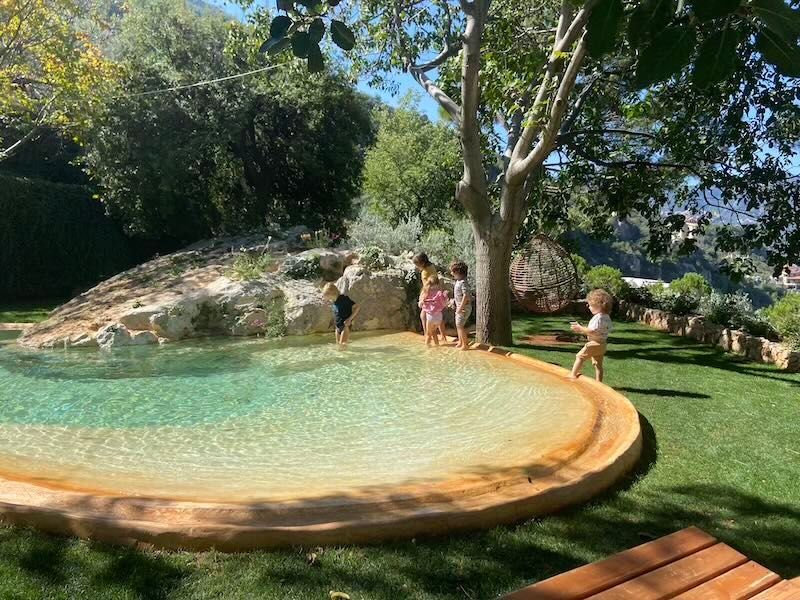Circular natural swimming pool with turquoise water surrounded by rocks and lush green lawn in a sunny outdoor setting.