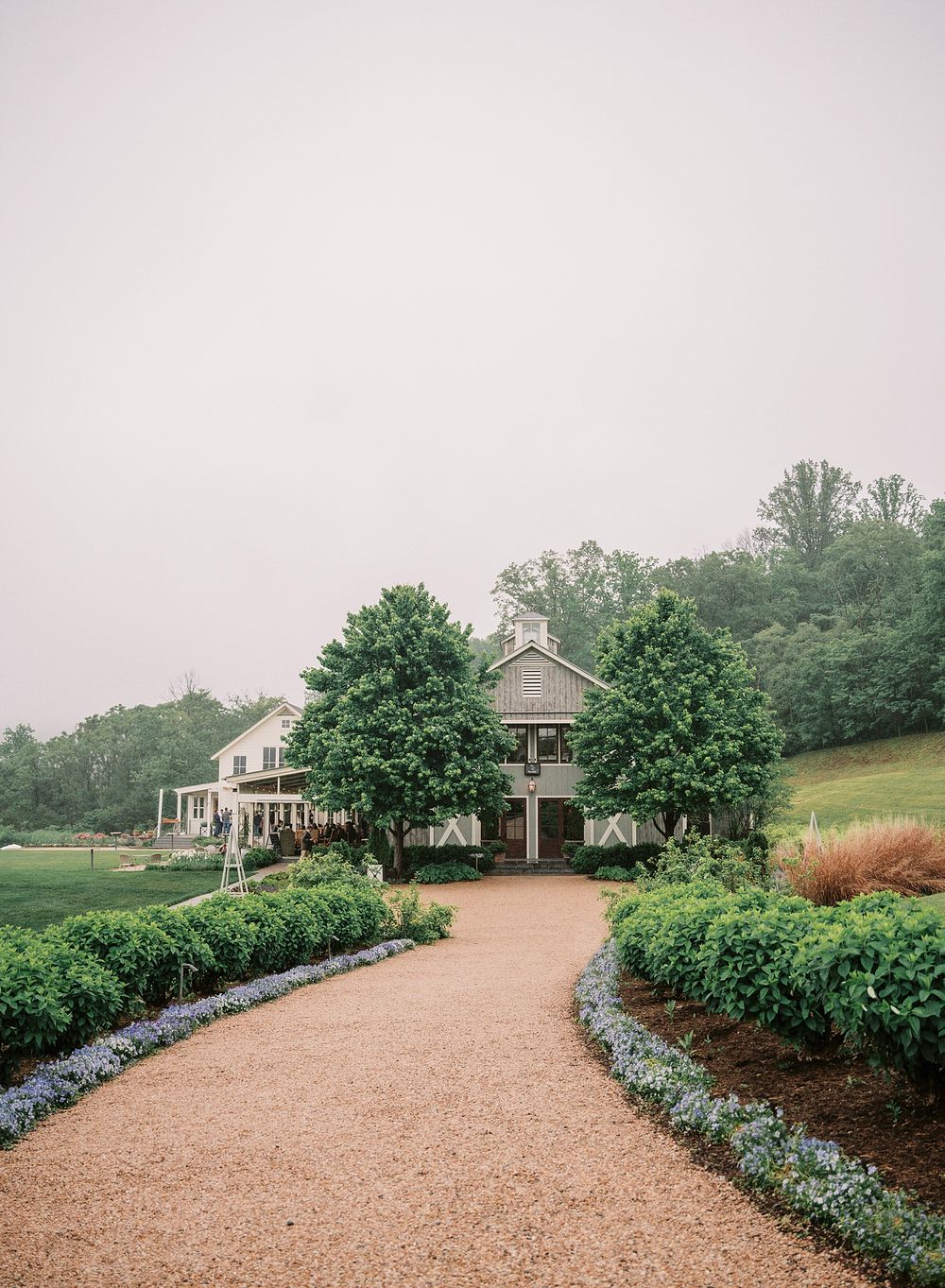 A grand country estate with a curved gravel driveway lined by manicured gardens and blue flowers leads to an elegant home.