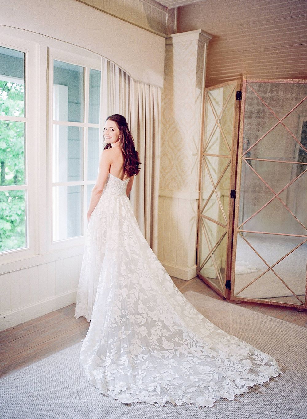 Elegant lace wedding dress with long train shown from behind in a sunlit room with white French windows.