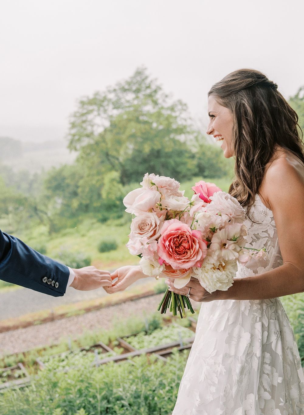 A person in a white lace wedding dress holding a beautiful bouquet of pink and blush roses reaches for someone's hand outdoors.