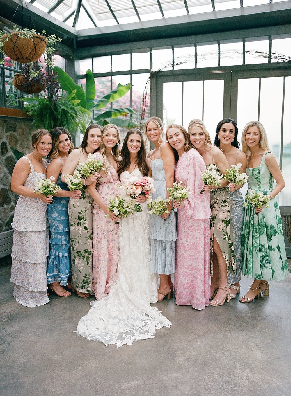 A wedding party in floral and pastel dresses poses in a greenhouse with elegant bouquets.