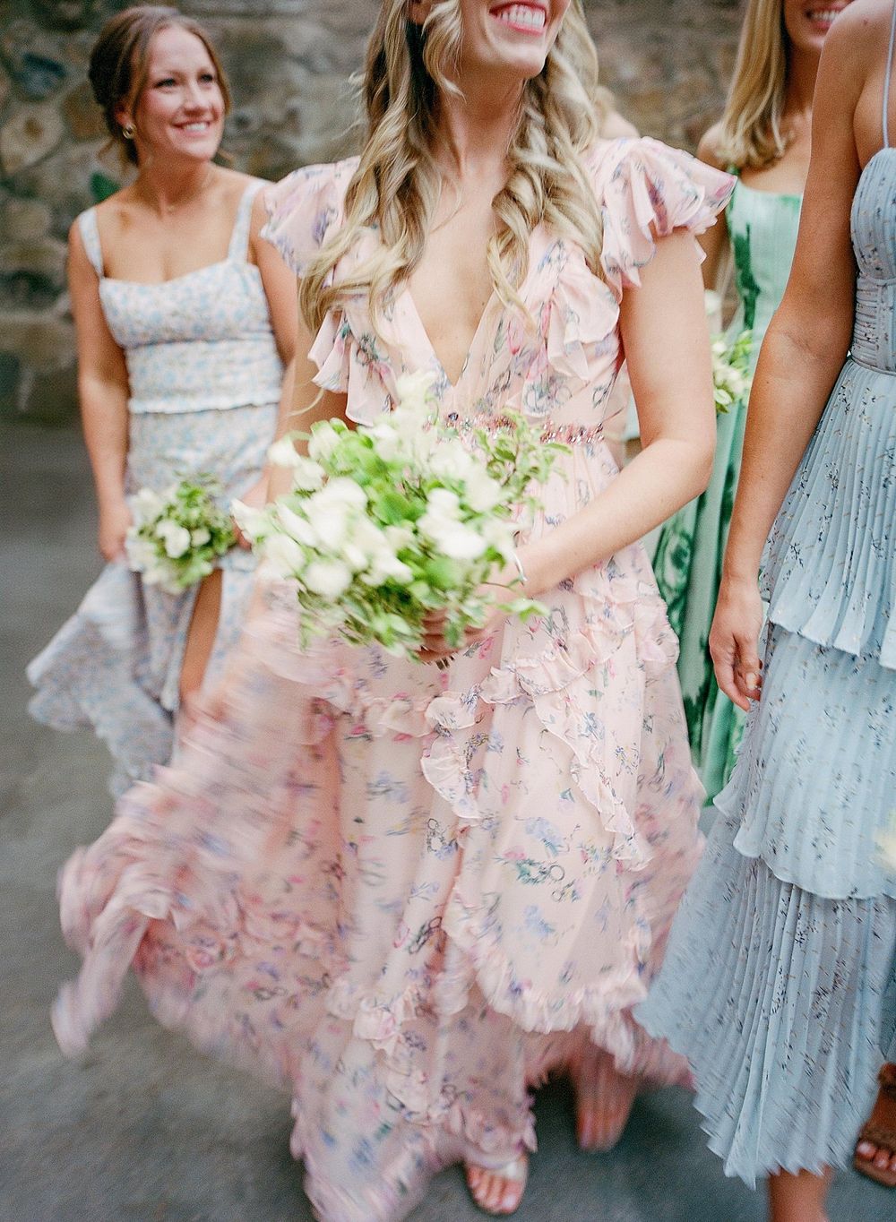 Bridesmaids in pastel dresses laugh and smile while holding white bouquets at an outdoor wedding celebration.