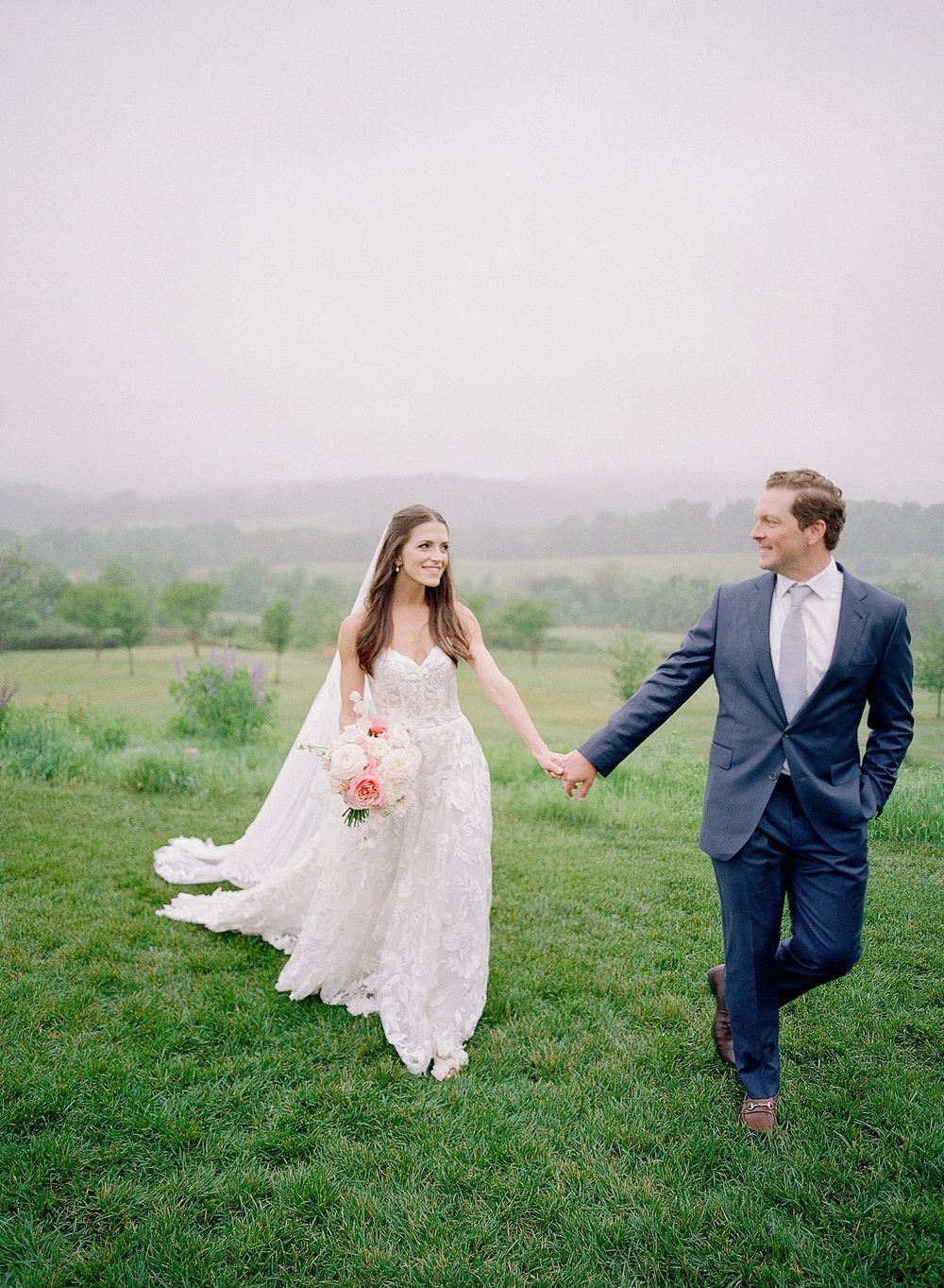 A bride and groom hold hands while walking across a misty hilltop meadow on their wedding day.
