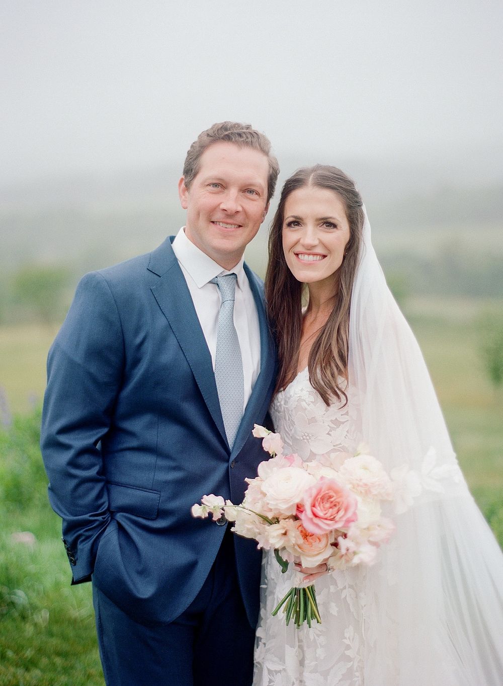 A joyful couple embraces on their wedding day outdoors with a misty landscape backdrop and pink bouquet.