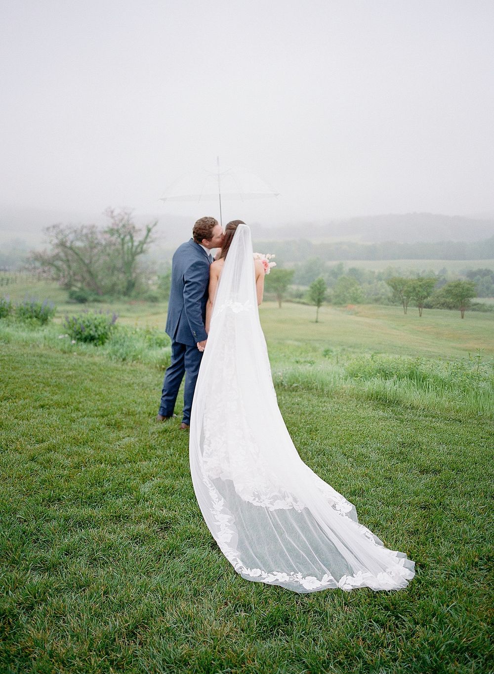 A couple shares an intimate moment on a misty hilltop, with a flowing white wedding veil trailing behind on green grass.