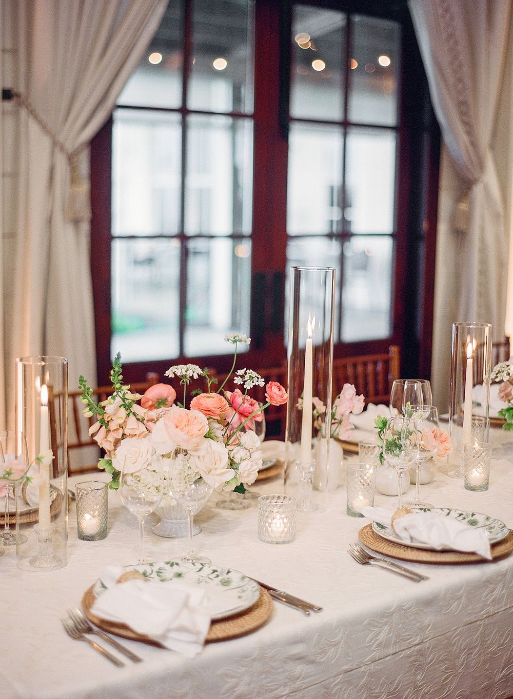 Elegant wedding table setting with coral and white flowers, gold chargers, and tall candlesticks by a window with cream curtains.