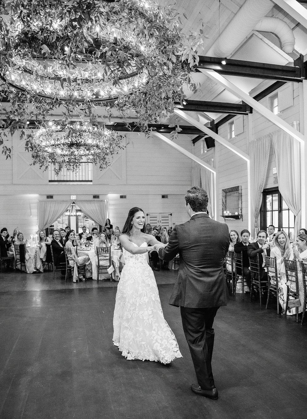 A couple shares their first dance in an elegant ballroom with chandeliers and florals while guests look on.