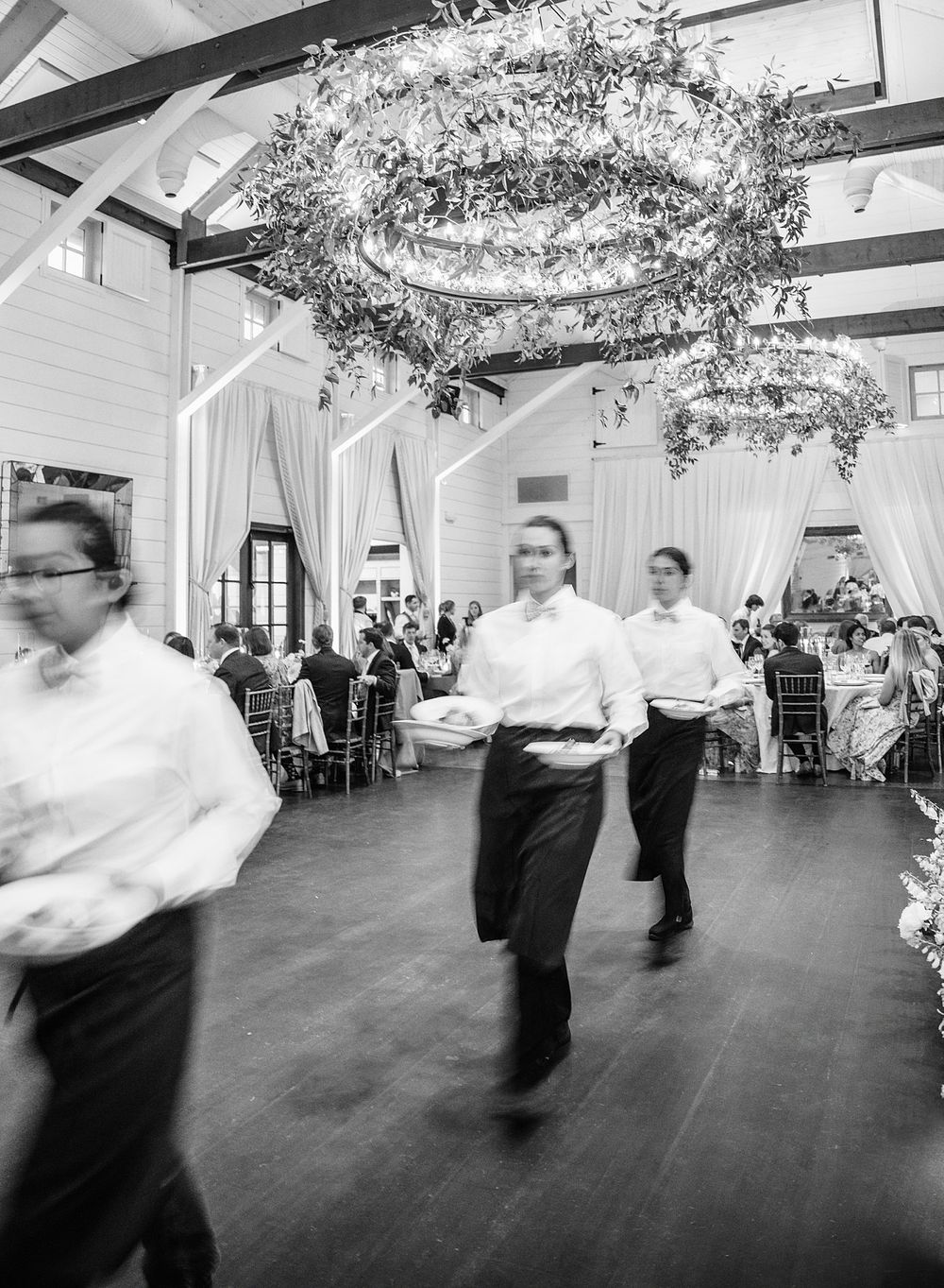 Waiters in white shirts move swiftly across a dance floor beneath an elegant floral chandelier during an indoor event.