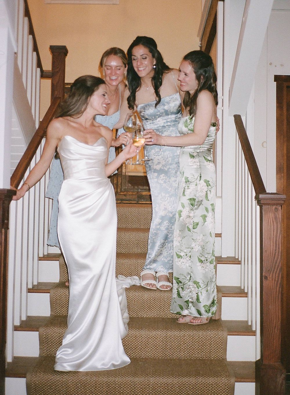 A group of women in formal dresses gather on carpeted stairs, with one wearing an elegant white satin gown.