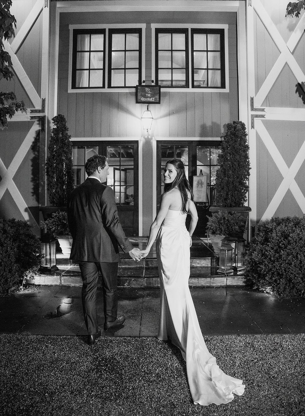 Black and white wedding photo of couple standing in elegant gown and tuxedo in front of chapel entrance at night.