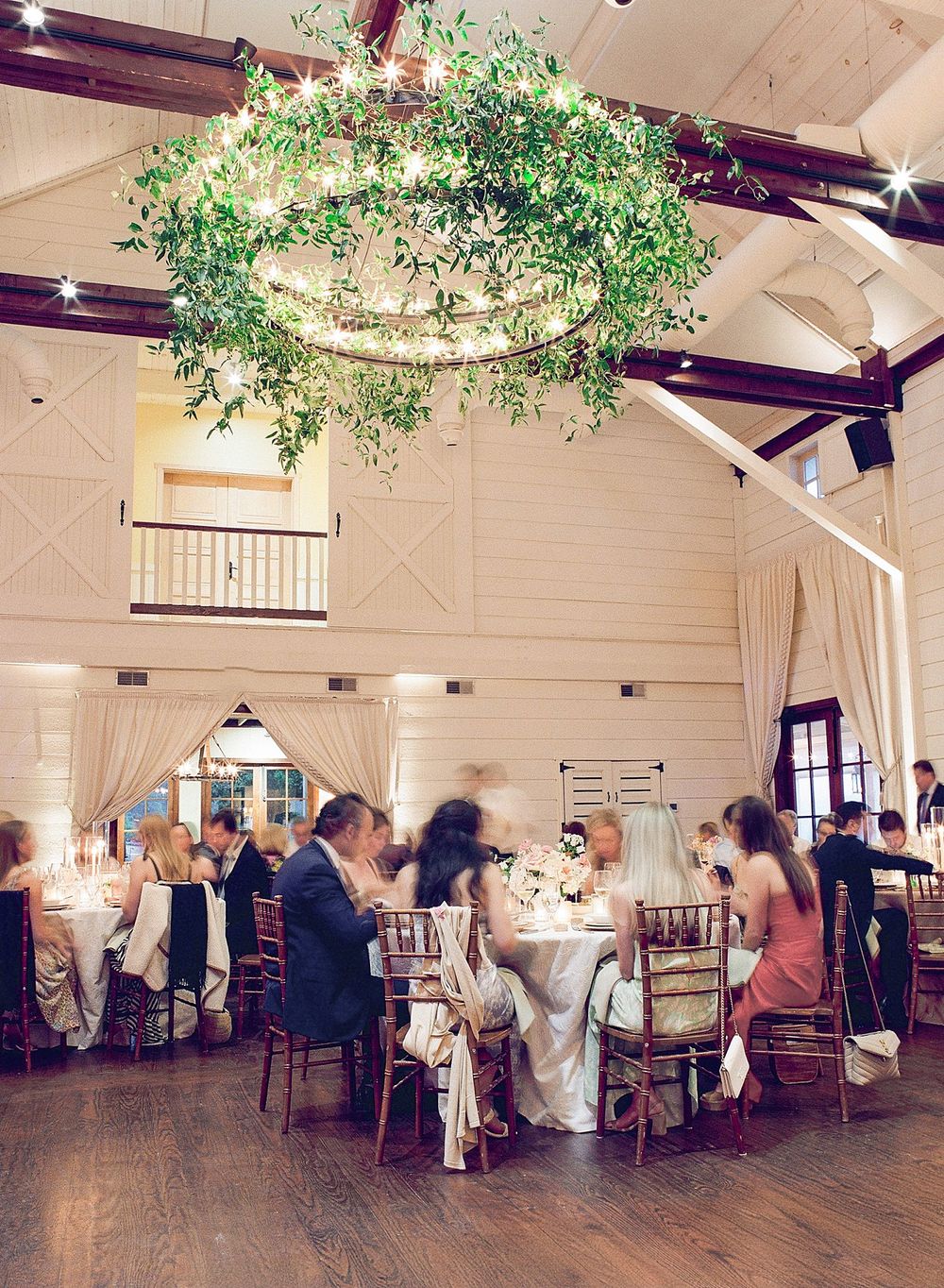 Elegant barn reception venue with high vaulted ceiling, hanging greenery chandelier, and round dining tables with guests.