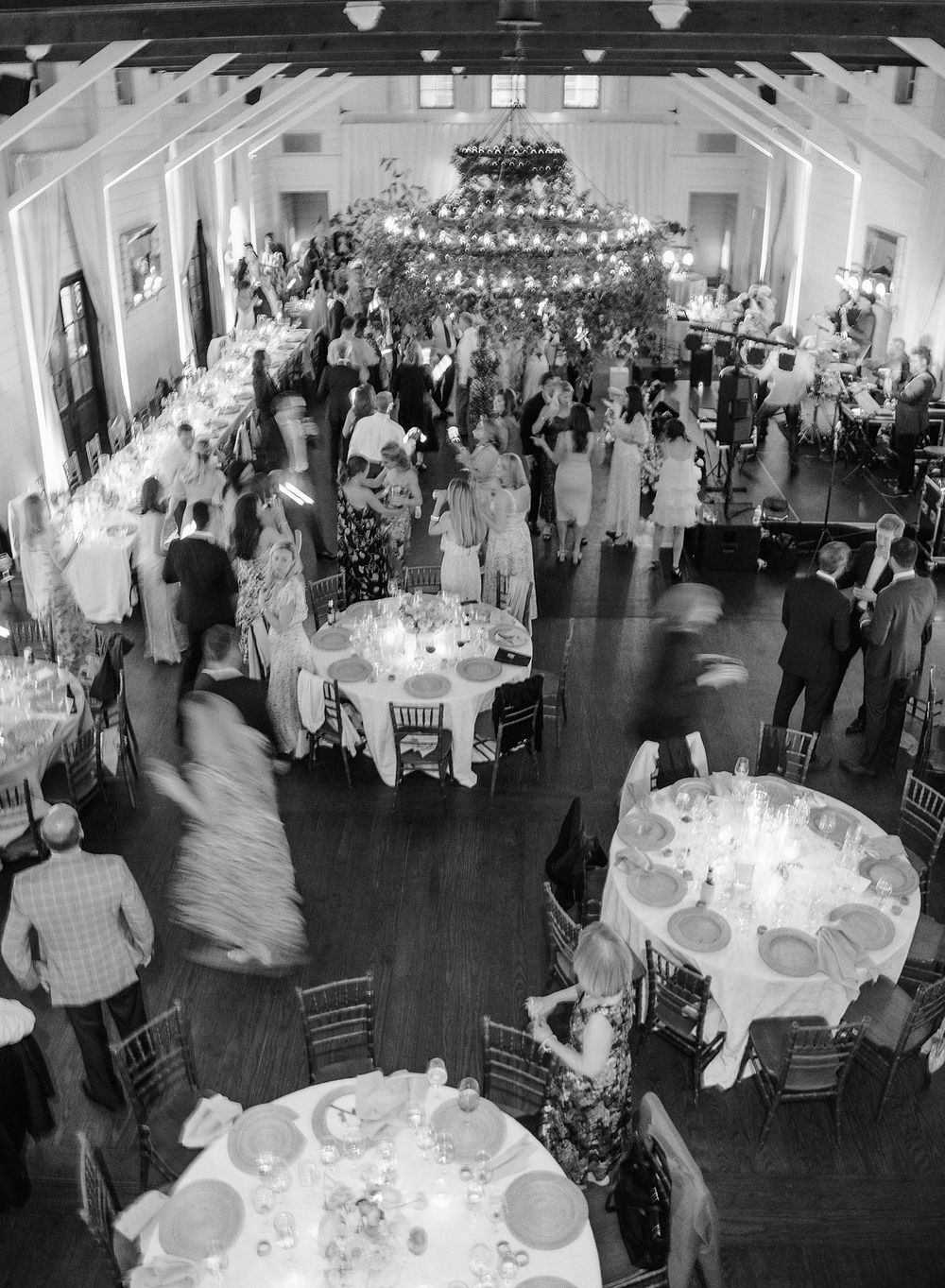 Black and white overhead view of an elegant wedding reception with round tables, chandeliers and guests dancing on a large dance floor.